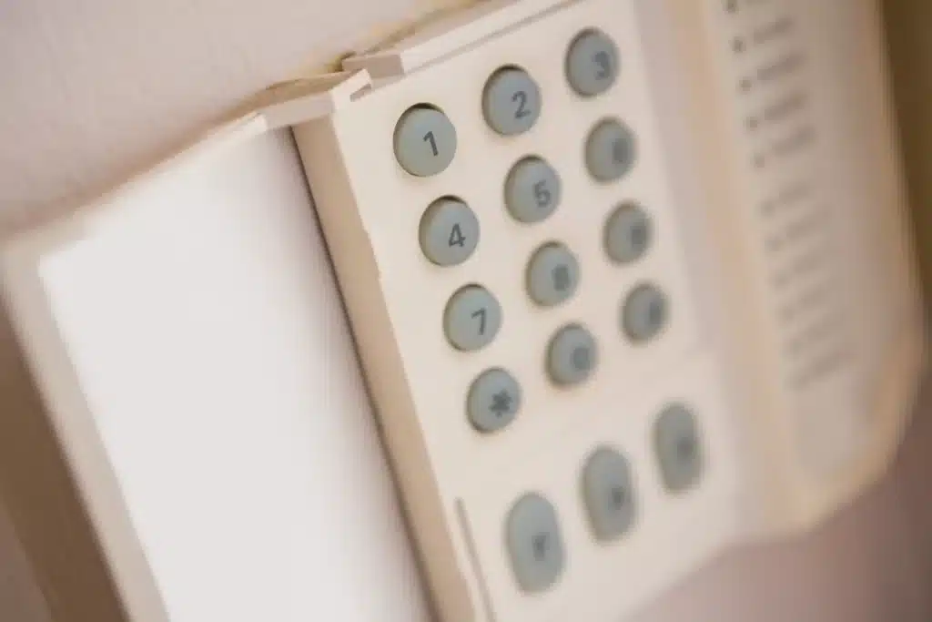 The image depicts a close-up view of a white, wall-mounted home security keypad with numbered buttons from 1 to 9, plus symbols, blurred in the foreground.