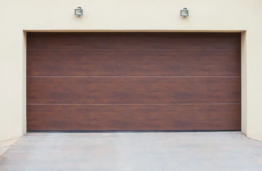 A large, closed, brown garage door set in a beige wall with two wall lamps on either side, and a paved driveway leading up to it.