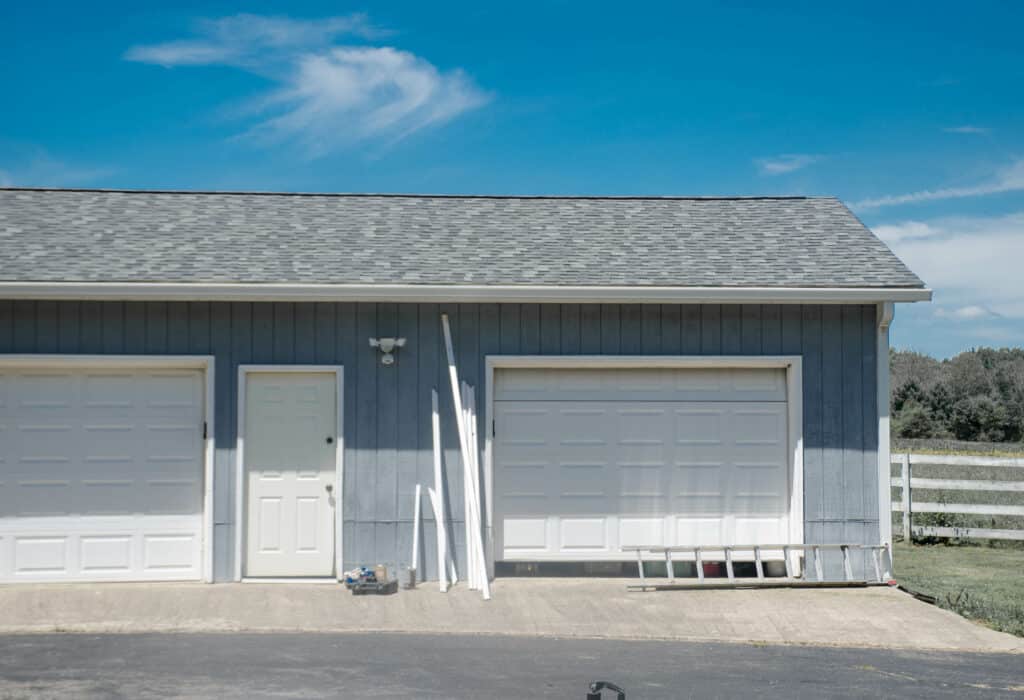 This image shows a grey building with two large garage doors and a single entry door. PVC pipes are leaning against the wall under a clear sky.