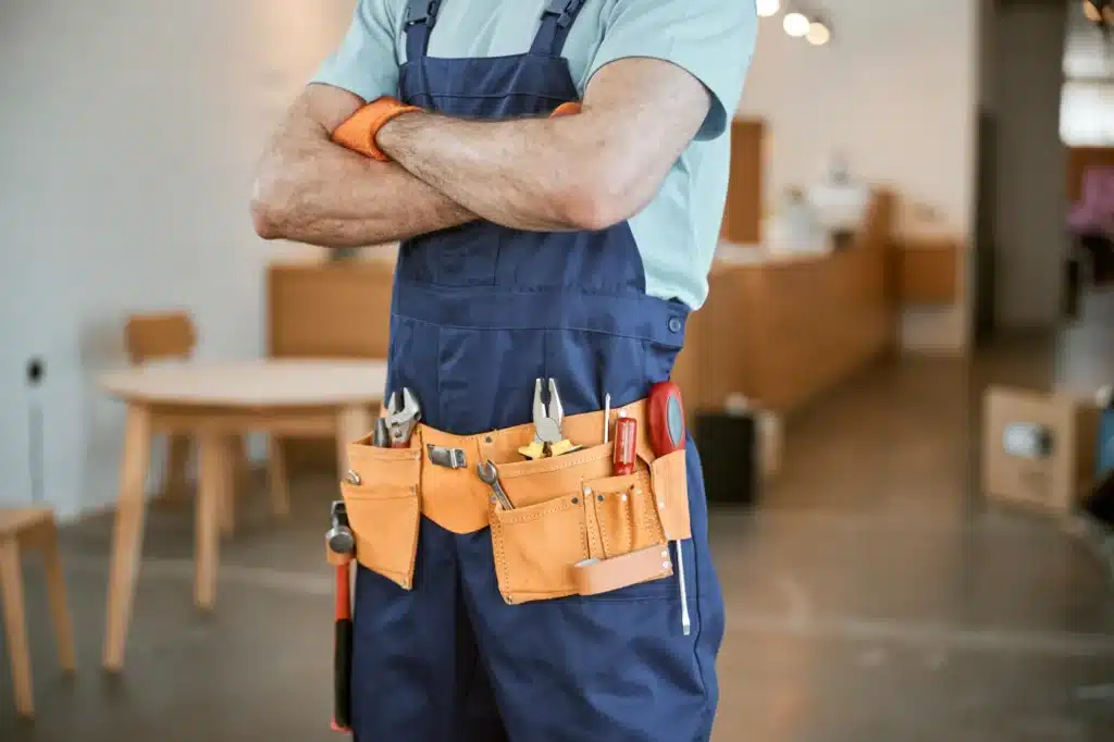 A person wearing a blue jumpsuit and an orange tool belt stands with arms crossed, showcasing various hand tools in a workshop environment.