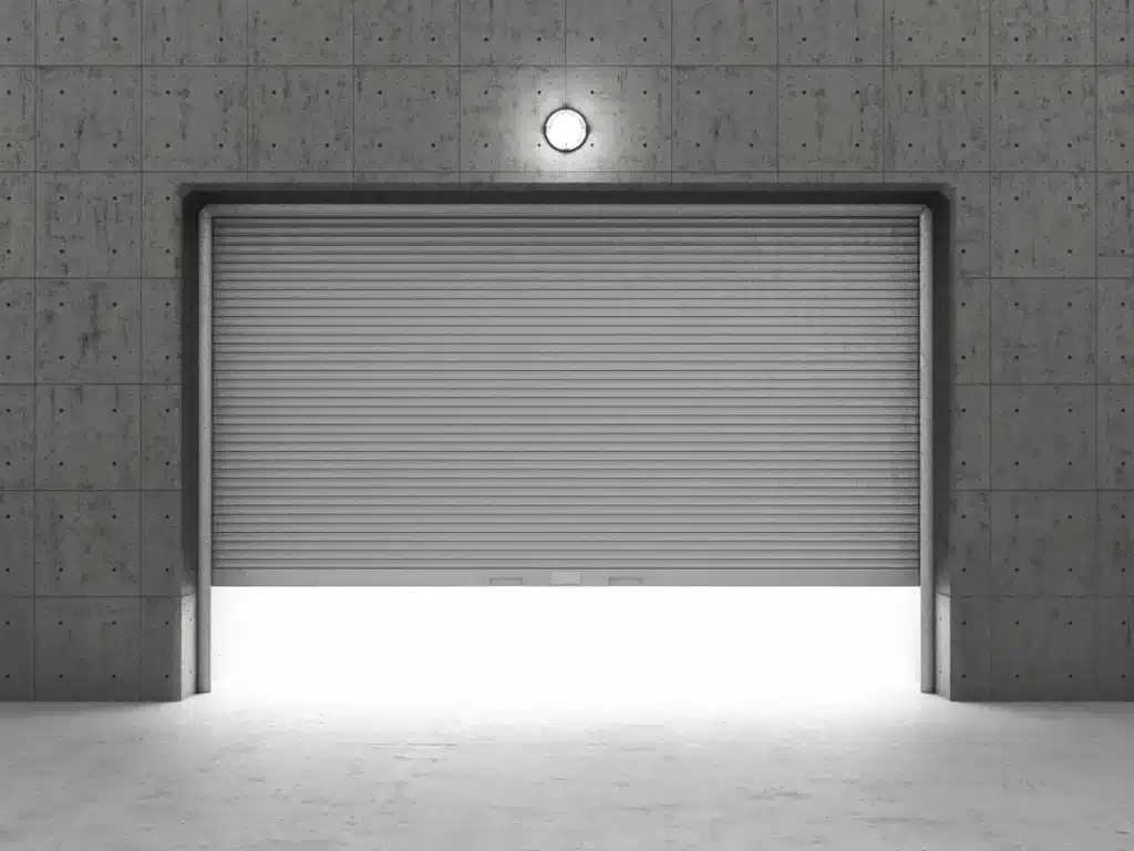 The image depicts a closed rolling shutter door in a minimalist concrete wall with a centrally mounted light above, casting a soft glow on the floor below.