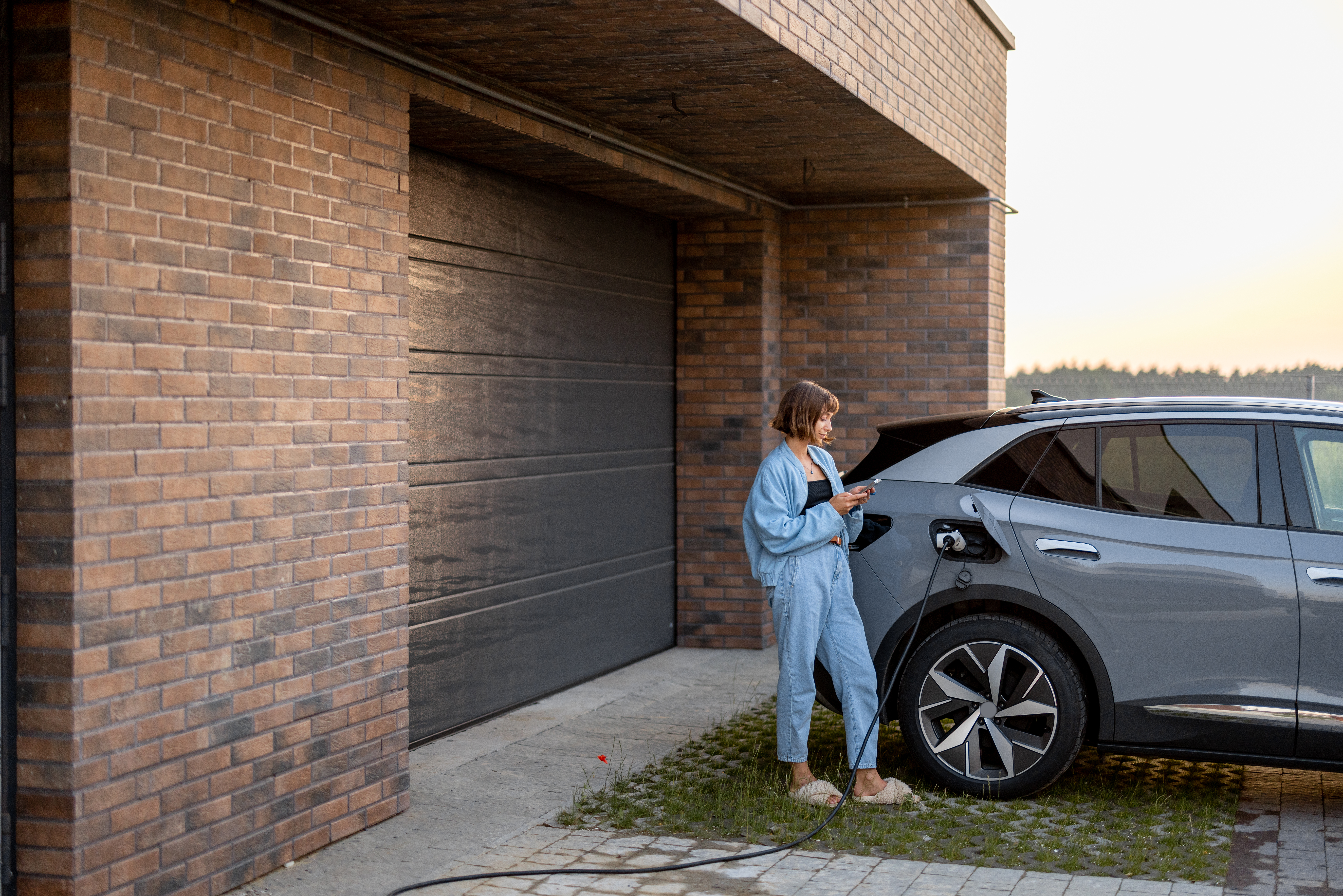 Young woman using smart phone while charging her electric car near garage of her house. Concept of modern lifestyle and sustainability