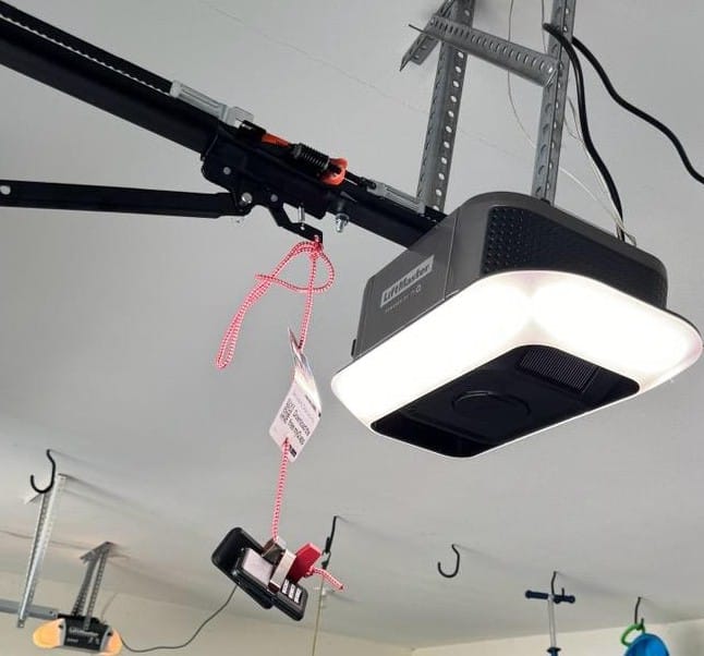 A garage door opener is mounted to the ceiling with a red and white emergency release handle hanging down on a cord. Bright light illuminates below.