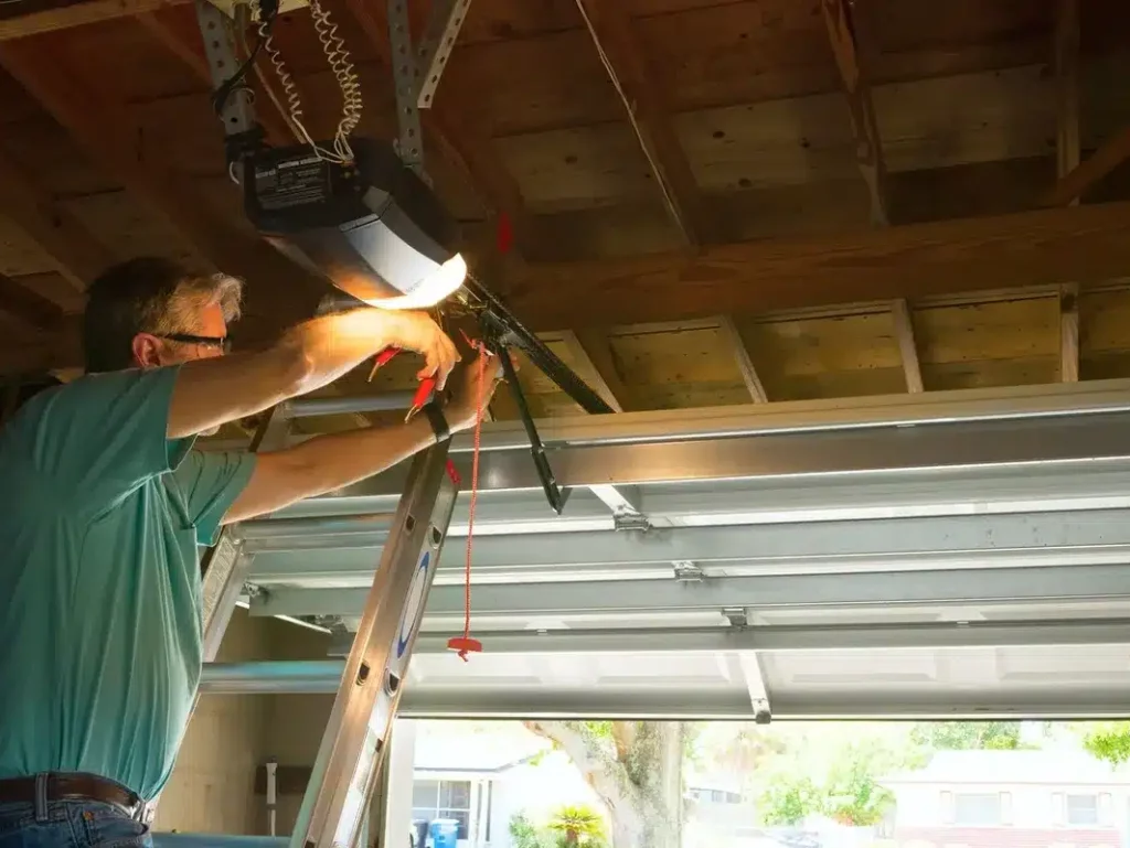 A person is on a ladder fixing a garage door opener. There's a wooden ceiling above and part of the garage door visible.