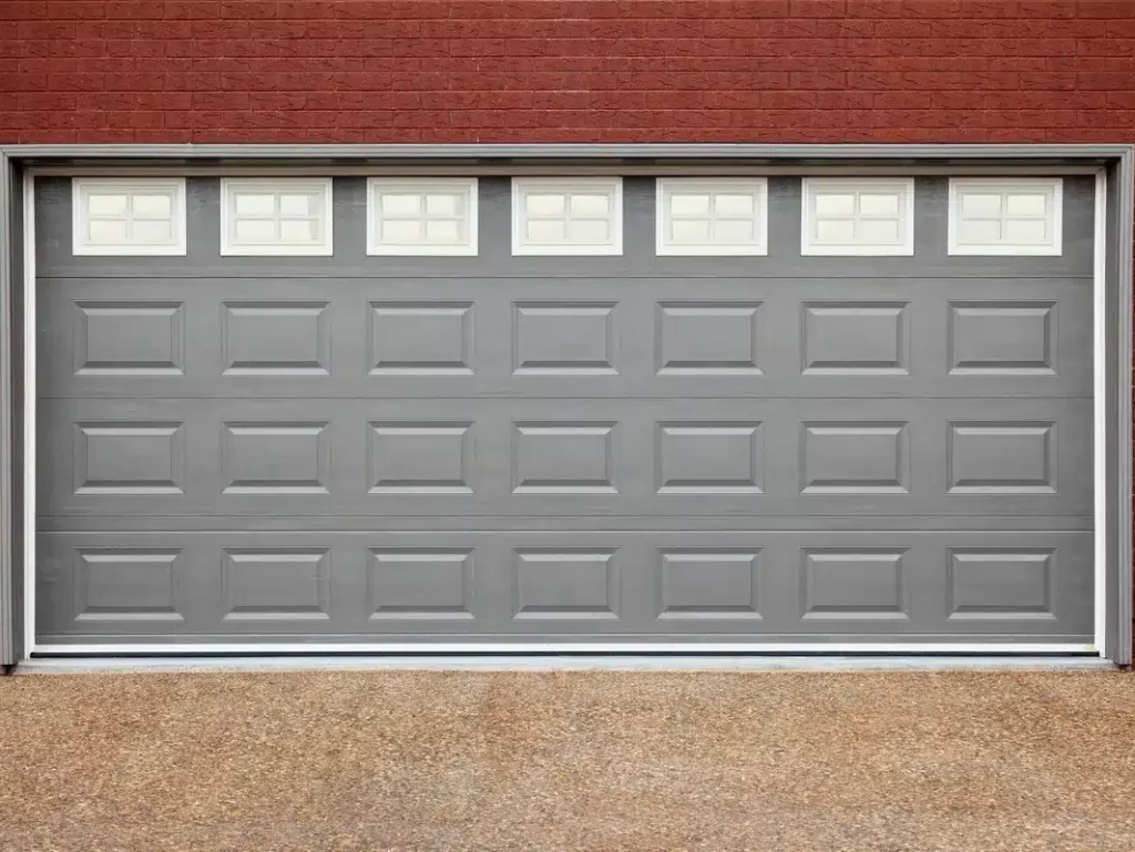 This is an image of a closed, gray, sectional garage door with a row of windows on a brick wall, flanked by two black outdoor lights.