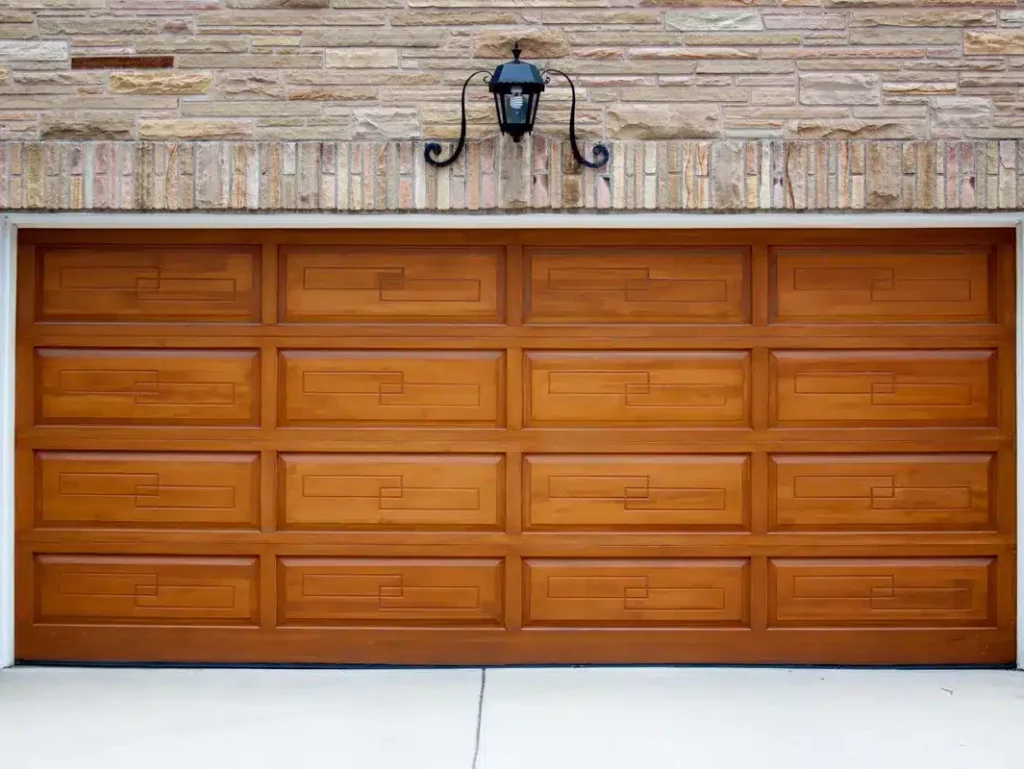 A large wooden garage door is shown with rectangular panels, set into a stone facade house, under a single black lantern-style wall light.