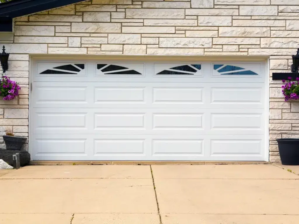 A residential double garage door made of white panels with decorative windows, flanked by hanging flower baskets and a wall-mounted lantern.